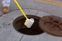 An employee collects a wastewater sample from an open manhole. A plastic bottle at the end of a long, yellow pole contains the water sample as it's lifted from the manhole.