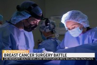 A still from a video of medical workers in surgical gowns and masks. Text on the screen reads, "Breast cancer surgery battle. CBS / 素人色情片Health News investigation into reconstruction costs.