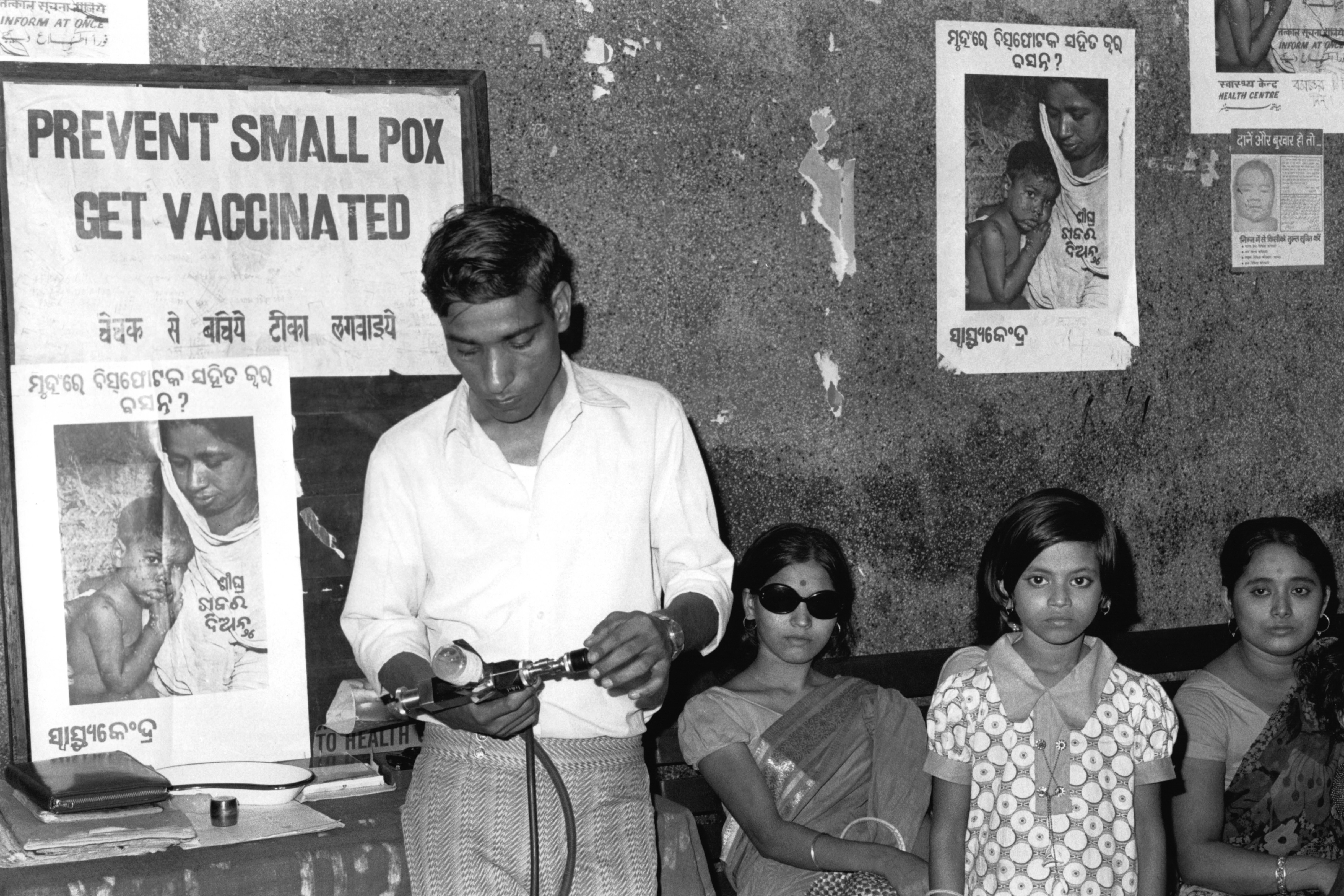 In this 1974, black and white film photograph, a doctor is standing beside two women and one young girl who are waiting to be vaccinated. He holds a jet injectorIn his hands, preparing for vaccination. Beside him and on the walls are posters that say, 鈥淧revent Smallpox 鈥� Get Vaccinated鈥� and show images of a mother holding a child with smallpox.