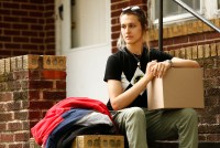 Hal Dempsey is photographed sitting on the brick steps of the house they are moving out of. A cardboard box is balanced on their knee, and a pile of clothes is to their right. They wear a T-shirt and cargo pants and are looking away from the camera.