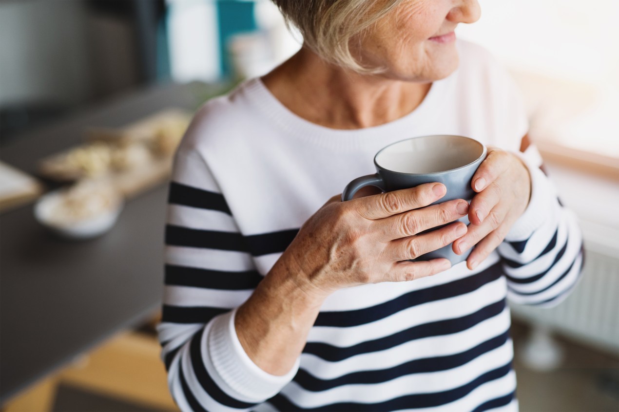 A photo of an older woman holding a mug inside her house.
