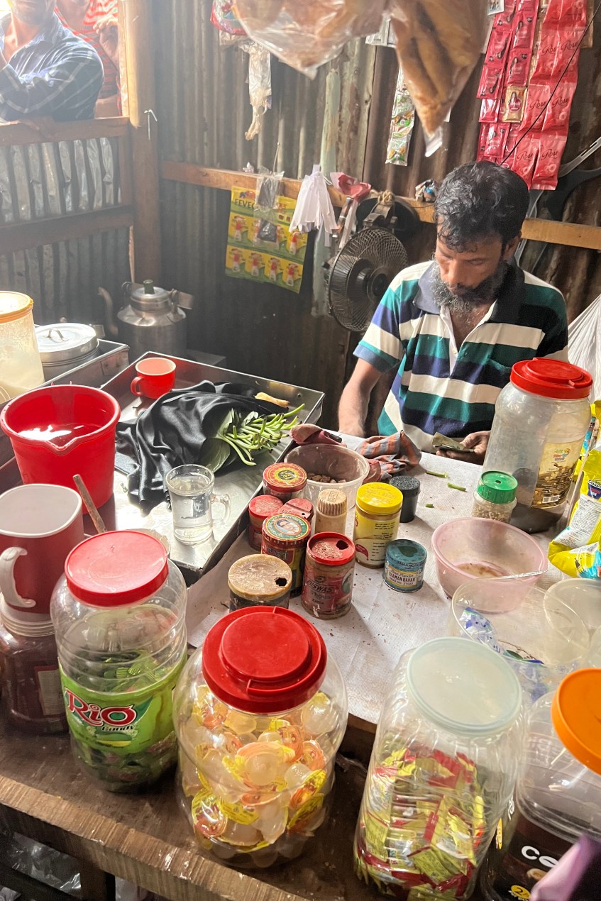 A man sits at a tea stall counter, the surface of which is covered in colorful canisters. Behind him and to his right are a tea kettle and a metal fan.