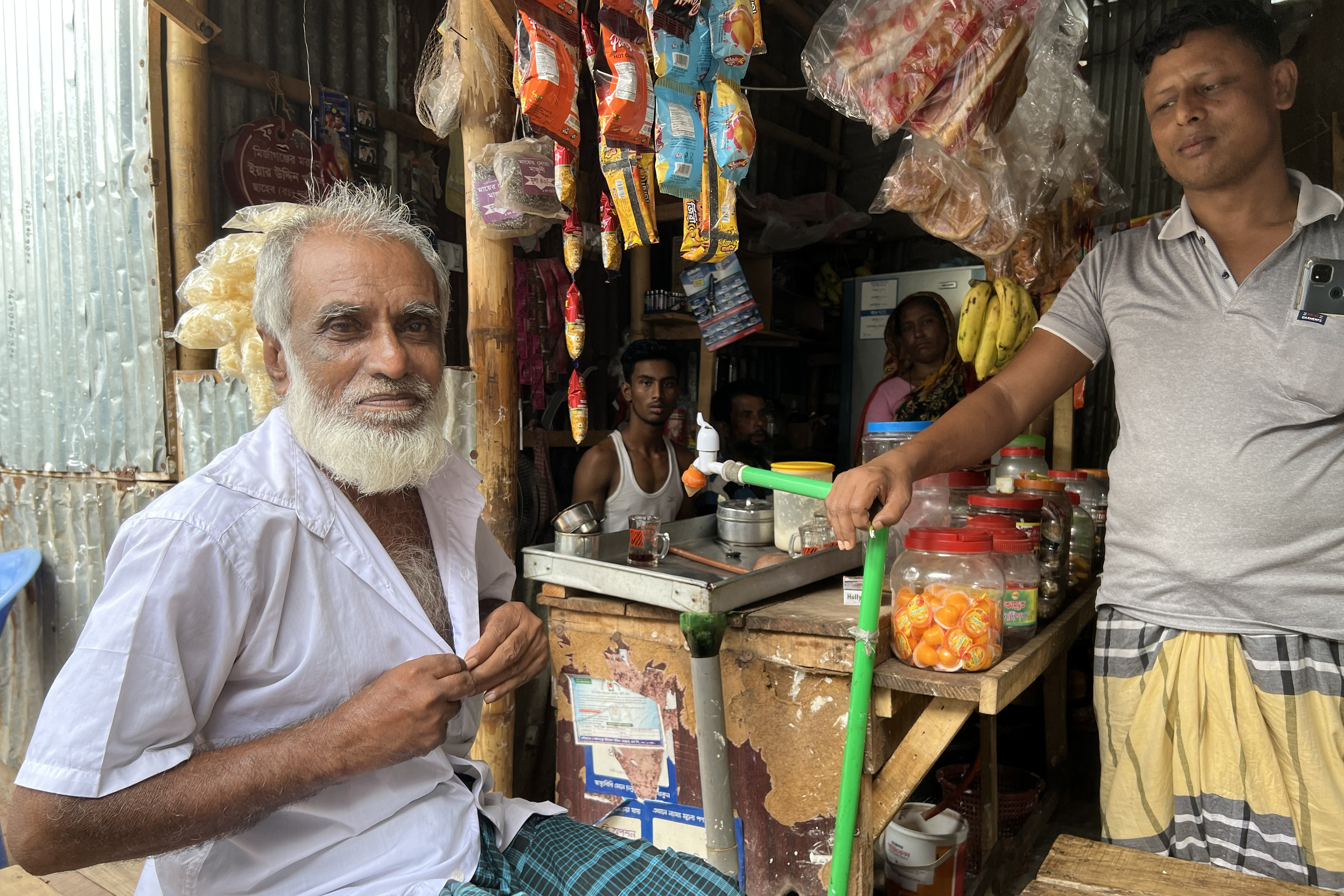 Shohrab, a man in his early 70s, sits outside of a tea stall on a well-worn wooden bench near his home in Dhaka. Inside the stall, a colorful display of snacks and sweets hang from the ceiling. Three other men and one woman are nearby in the tea stall.
