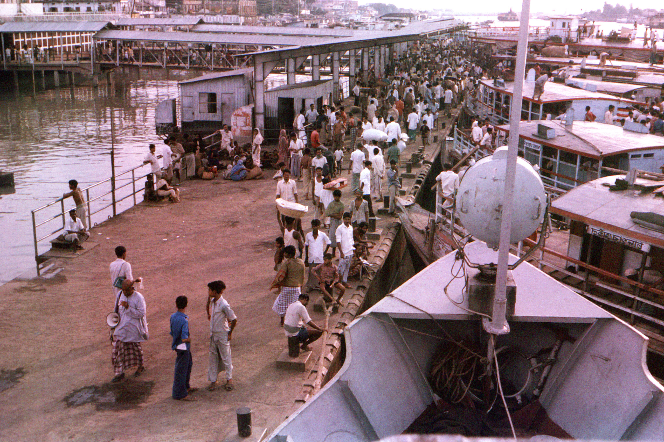 An overhead view of a ferry terminal in Dhaka, Bangladesh, in 1975. A large crowd of people fill the main walkway, flanked by a row of boats on the right.