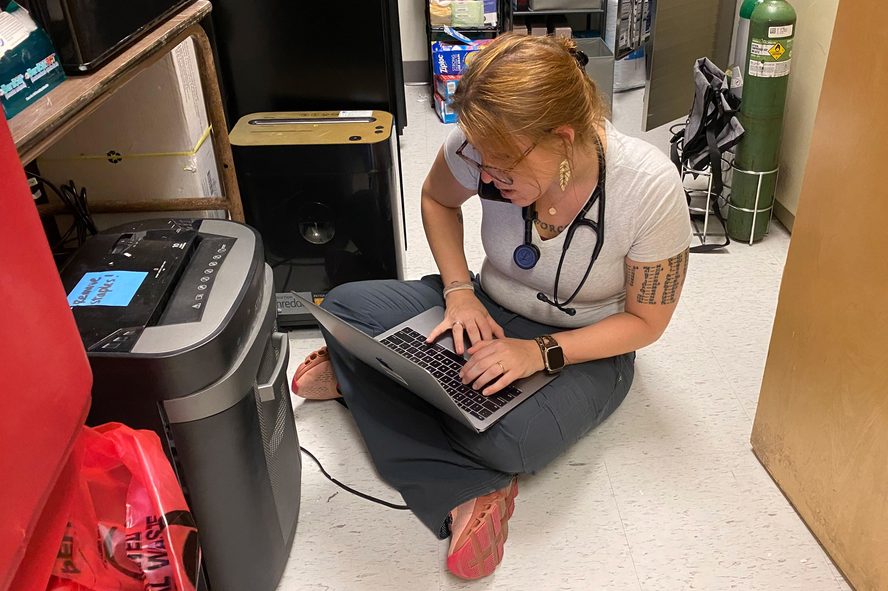 A photo of a doctor speaking on the phone while using her laptop. She is sitting on the floor.