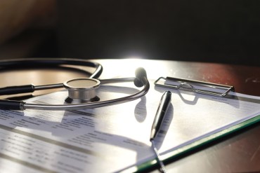 Medical documents on a clipboard are resting on a table or desk with a stethoscope and pen in sunlight.