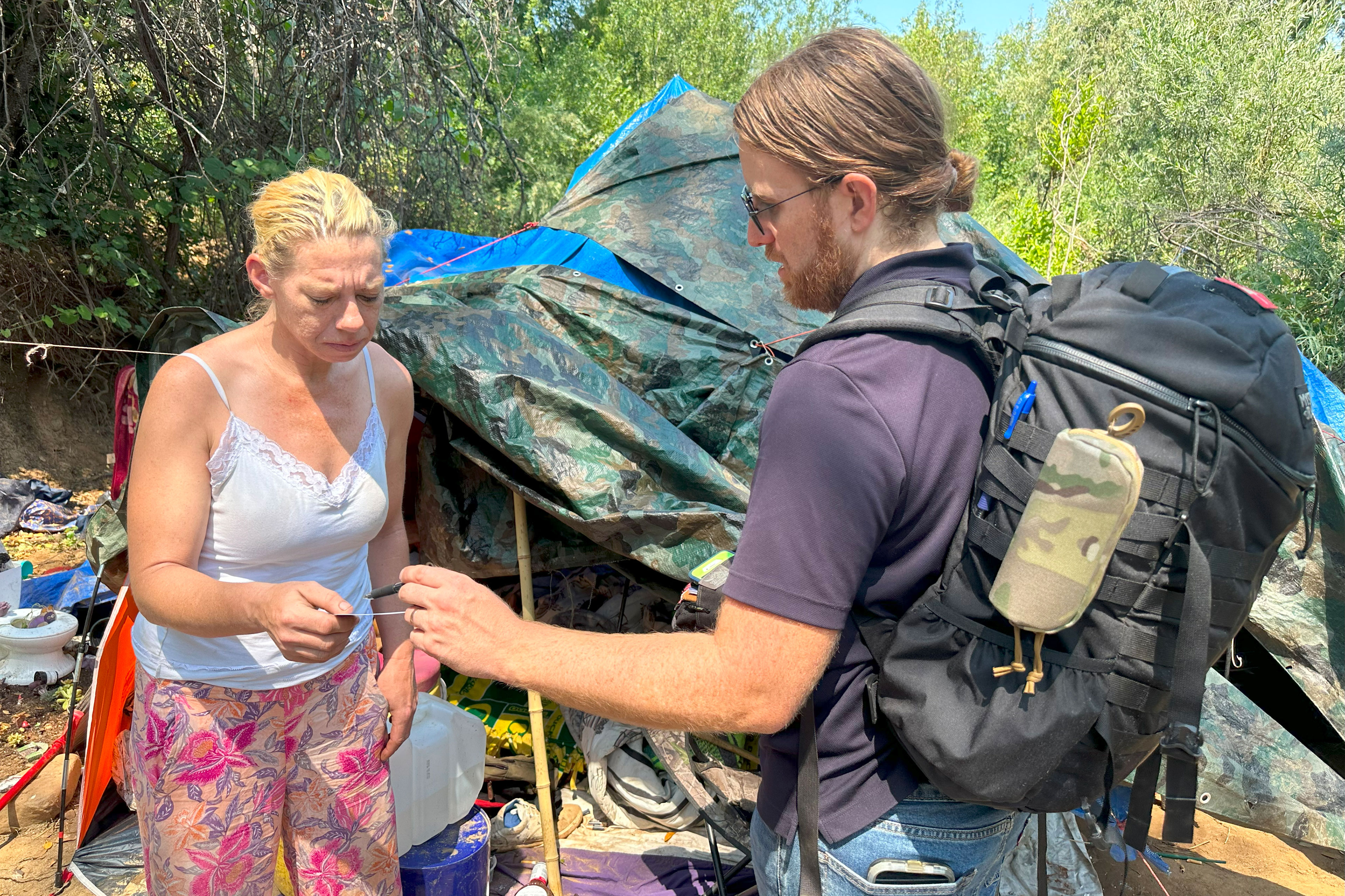 A man with long hair tied into a bun and wearing a black backpack shows something to a woman wearing a white tank top and pants with a floral pattern. They stand in front of a tent.