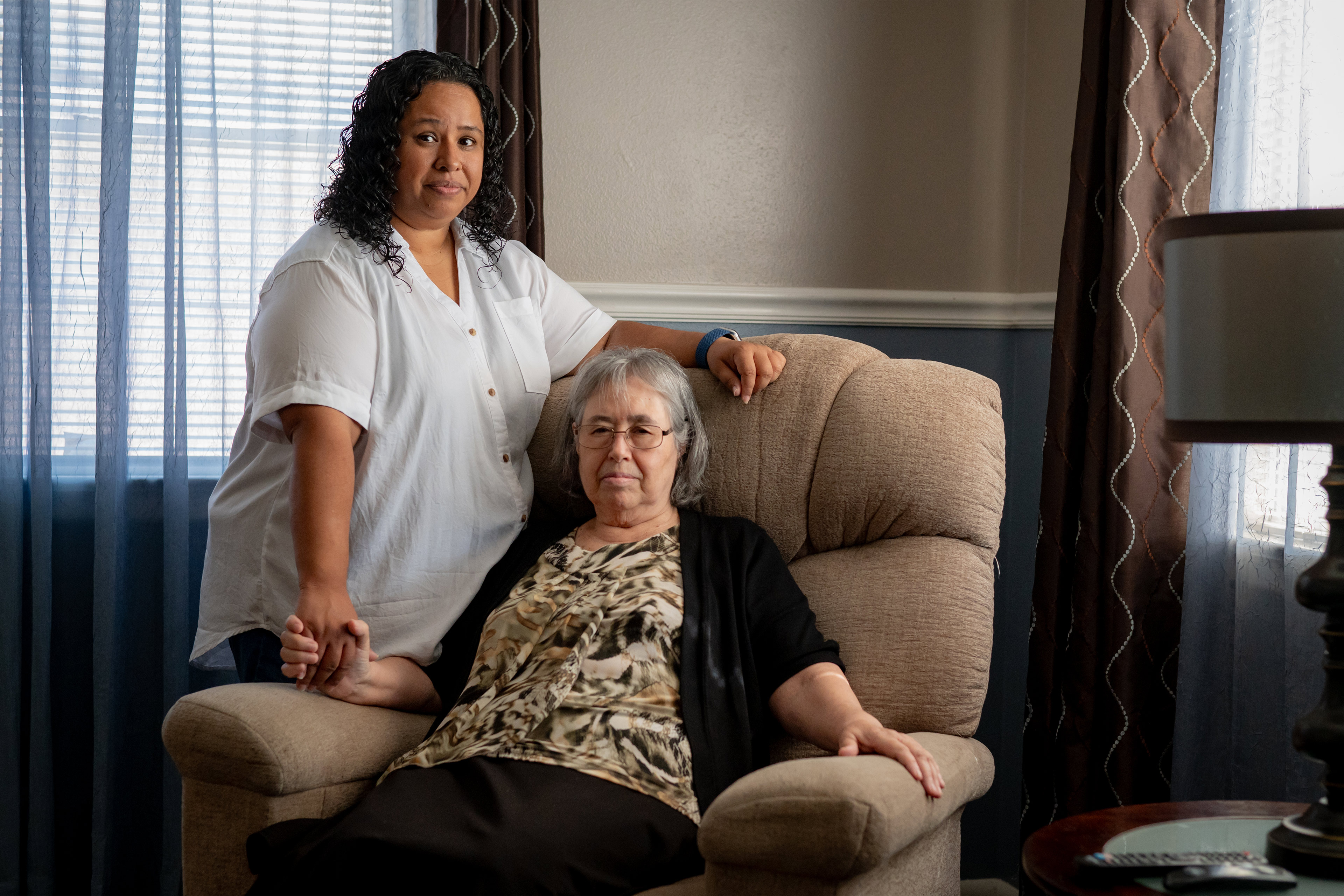 A photo of an older woman sitting in a chair while her adult daughter stands next to her.