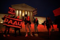 Members of the Progressive Anti-Abortion Uprising rally in front of the U.S. Supreme Court. They are illuminated by a strong red light. A woman standing in the center, while protesters around her hold signs with anti-abortion slogans.