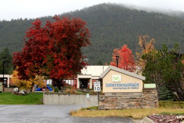 A yellow sign with blue lettering says "Intermountain, restoring hope for children" outside a building in Helena, Montana. A bright red tree and a playground can be seen in the background next to the building.