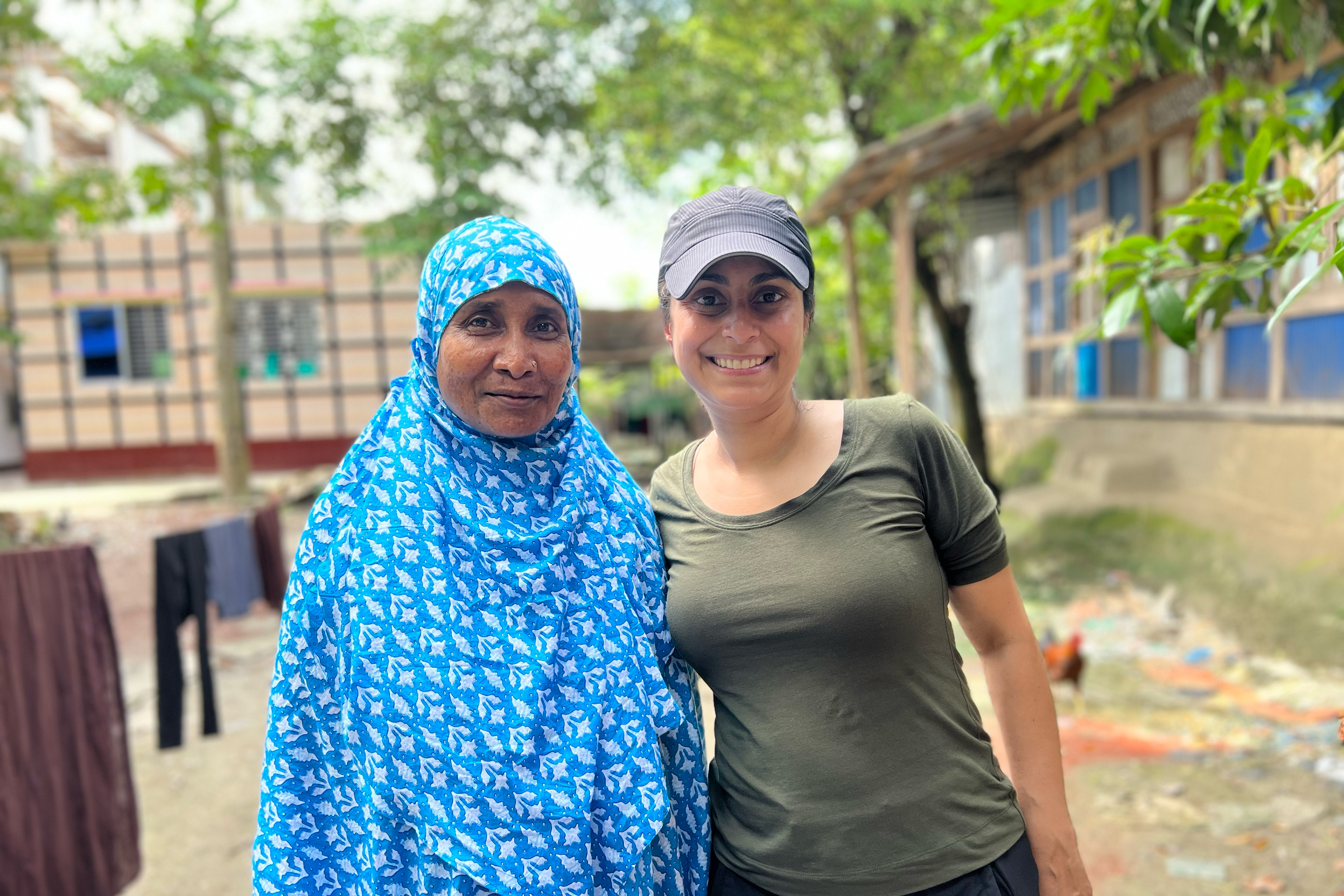 Rahima Banu (left) wears a blue scarf patterned with white flowers. It is draped over her head and body and tucked under her chin. She stands beside C茅line Gounder (right), who wears a black baseball cap and green T-shirt. They are in a small courtyard just outside Banu鈥檚 home. Laundry dries on a line in the background. Bright green, waxy leaves from growing trees provide shade and softened sunlight.