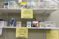 Donated prescription drugs are stacked on shelves. On the top shelf, a sign reads, "DONATED MEDICATIONS / INJECTABLES" On the shelf below, a second sign says, "DONATED MEDICATIONS / INHALERS / NASAL SPRAYS"
