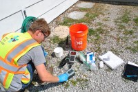 A person crouches on the ground as they test a water sample for PFAS. Various equipment is on the ground around them.