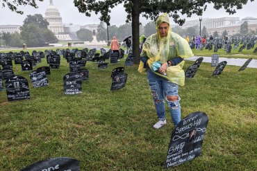 A woman wearing a transparent yellow rain jacket looks down at a sign stuck into the ground. There are dozens of similar markers in the background and the U.S. Capitol farther in the distance.