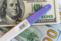 A positive pregnancy test rests on top of multiple $100 bills.